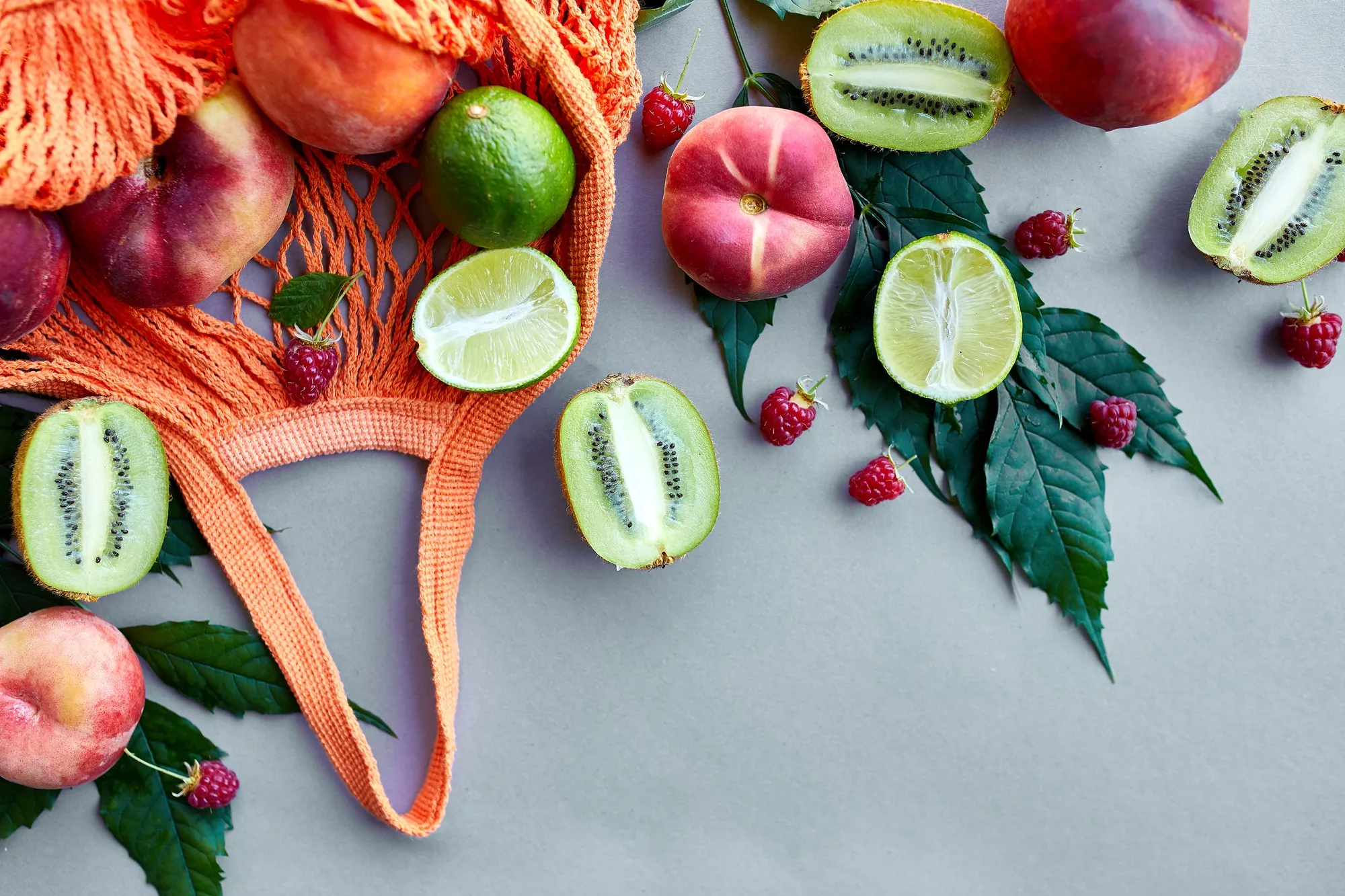 Flat lay of eco friendly mesh shopping bag  With Fruits raspberry, peach, kiwi, lime on grey surface in sunlight, summer time. Grocery concept, copy space, top view.