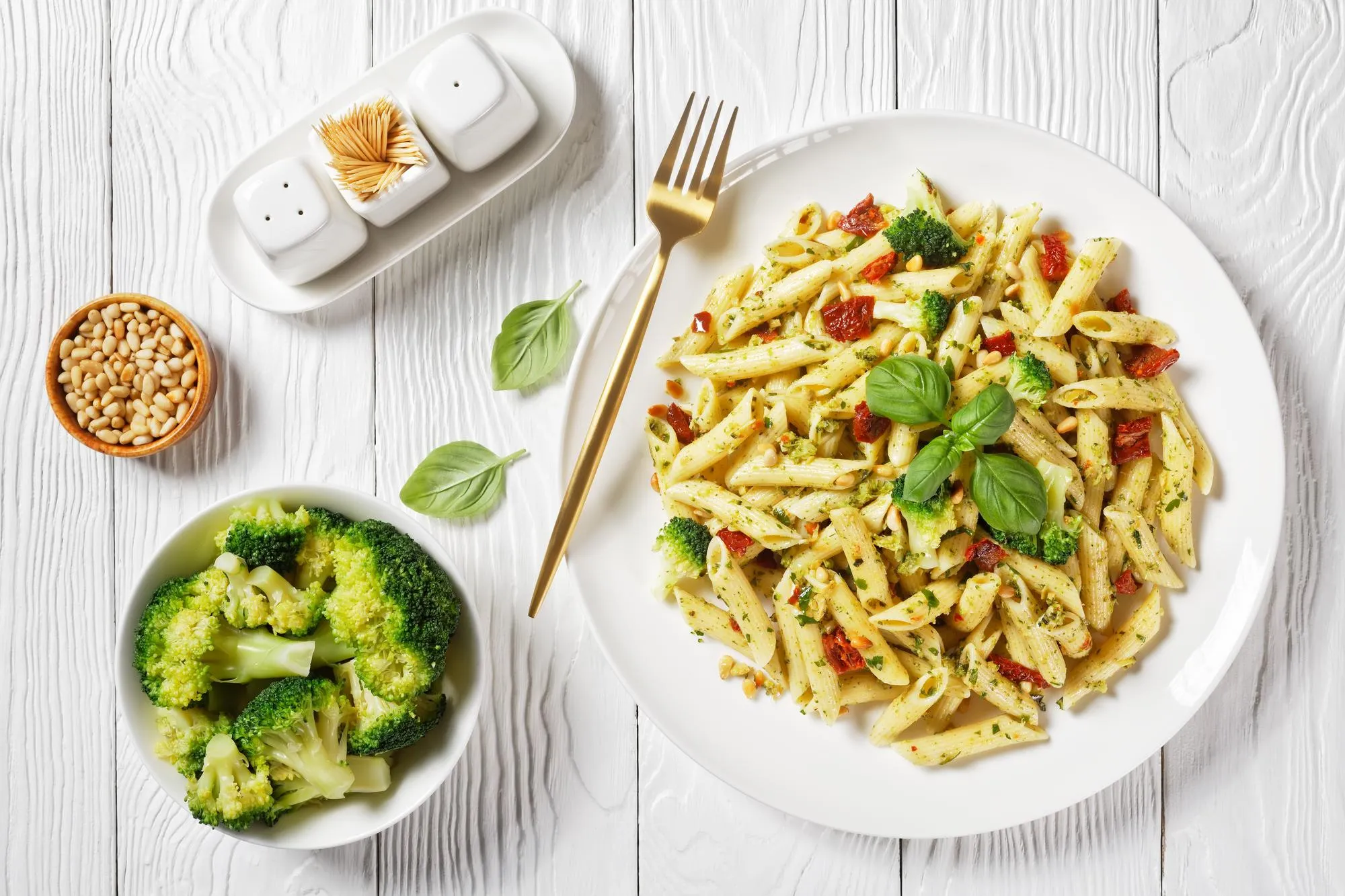 Broccoli pesto pasta with sundried tomatoes and pine nuts on a plate and ingredients on a white wooden table, horizontal view from above, flat lay