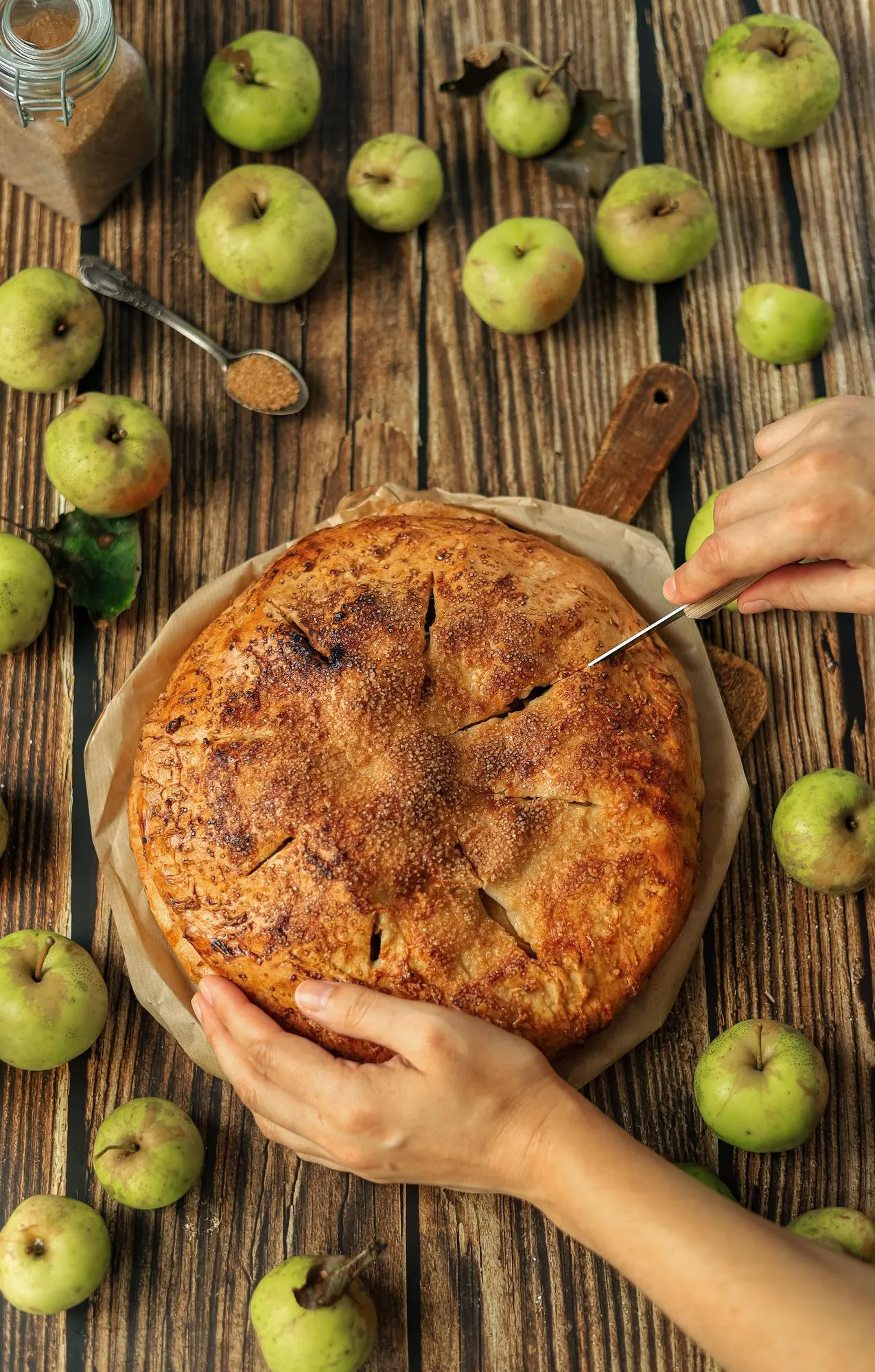 The chef's hands hold a fruit pie and cut a slice from it with a knife on a rustic kitchen table