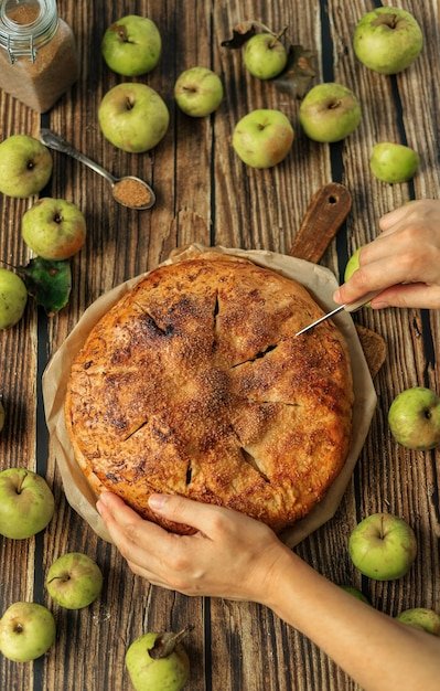 The chef's hands hold a fruit pie and cut a slice from it with a knife on a rustic kitchen table