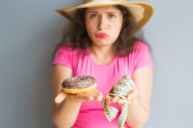 Confused young woman holding a donut and a measuring tape. Concept of Sweets, Unhealthy Junk Food and obesity.