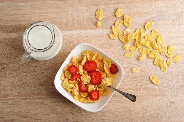 Cornflakes with strawberries and a jug of milk on wooden surface