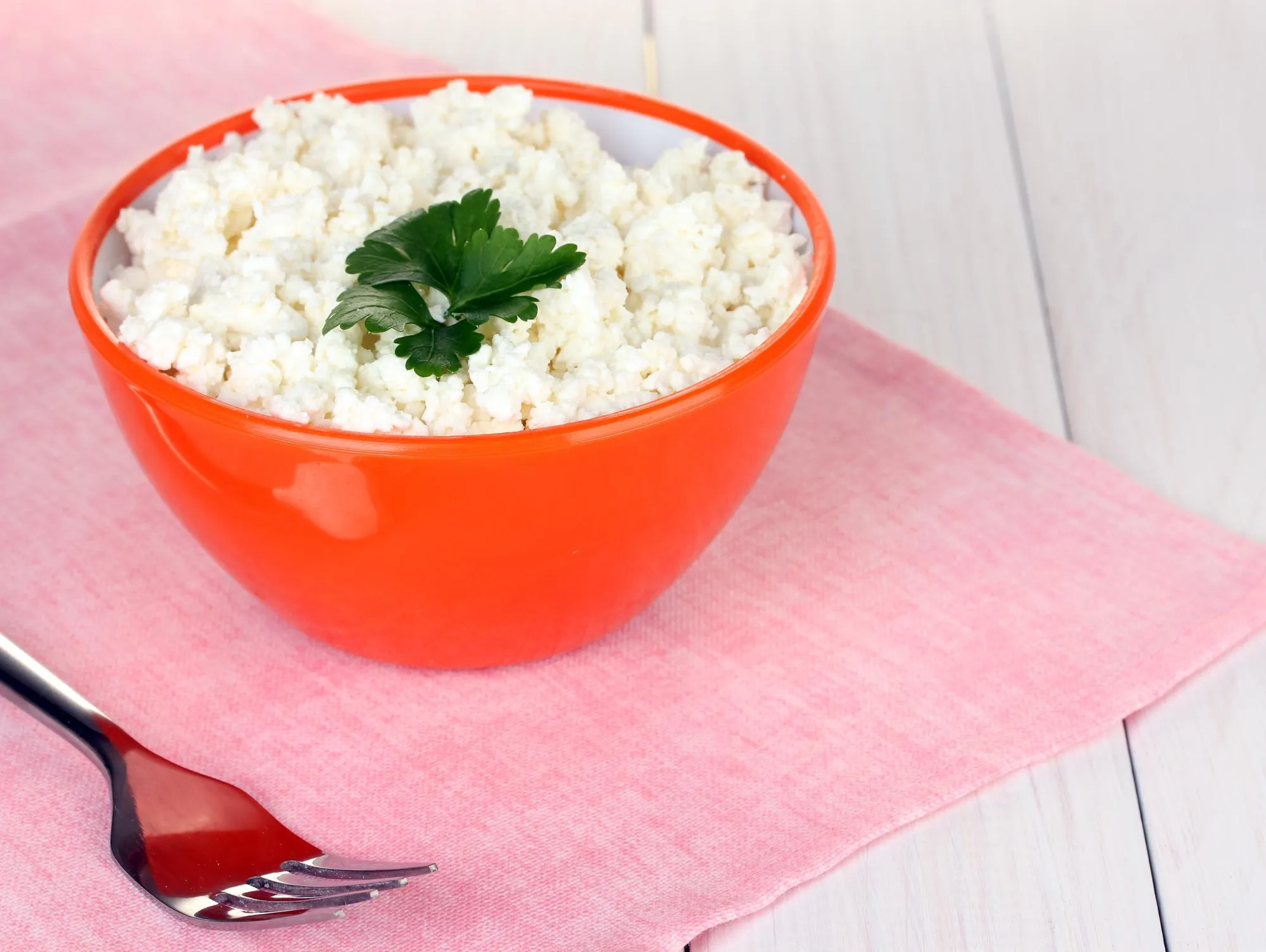 Cottage cheese with parsley in orange bowl and fork on pink napkin on white wooden table closeup