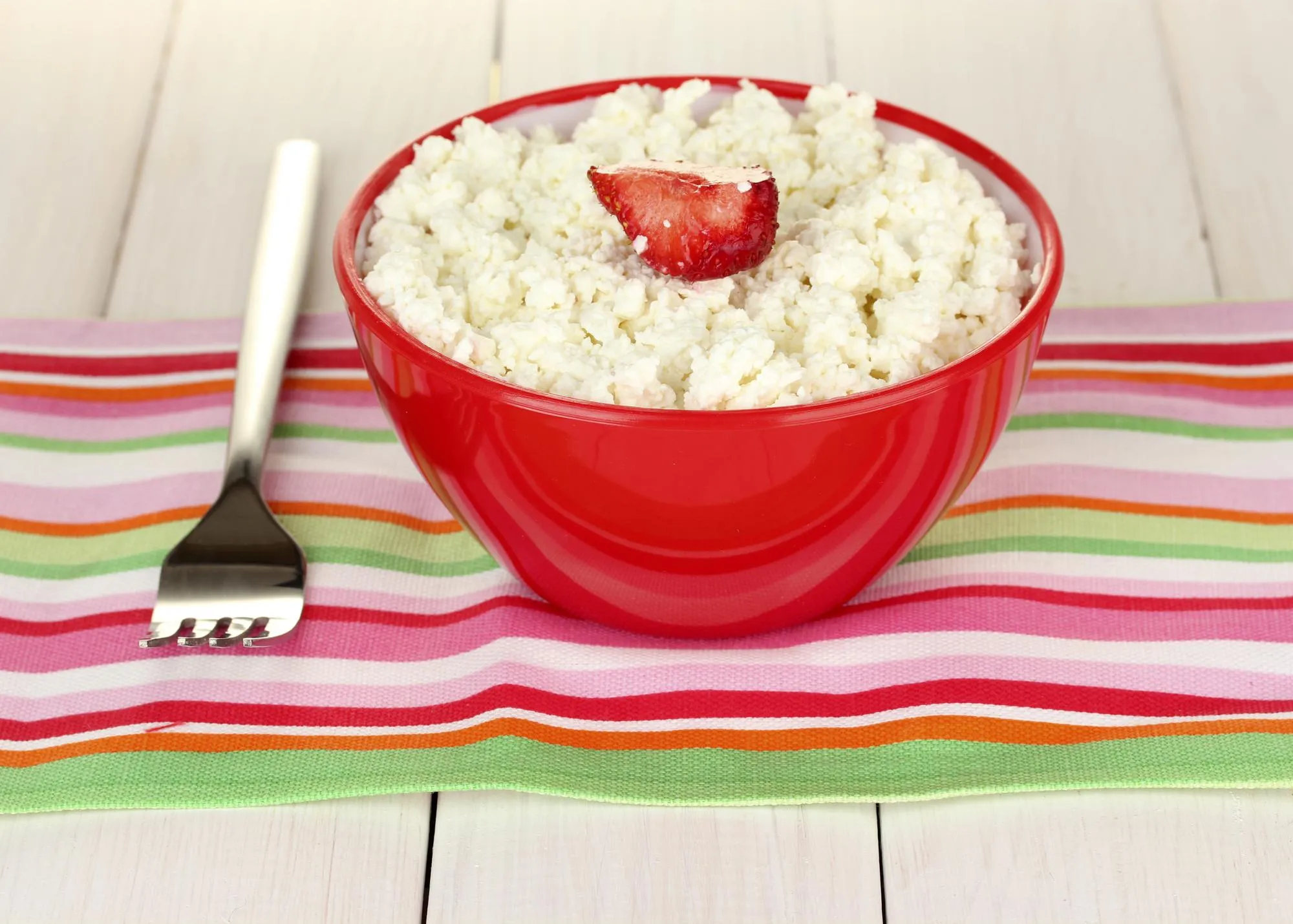 cottage cheese with strawberry in red bowl and fork on colorful napkin on white wooden table closeup