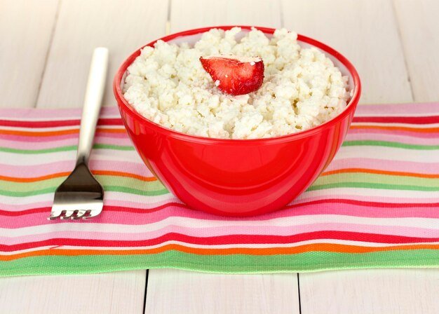 cottage cheese with strawberry in red bowl and fork on colorful napkin on white wooden table closeup
