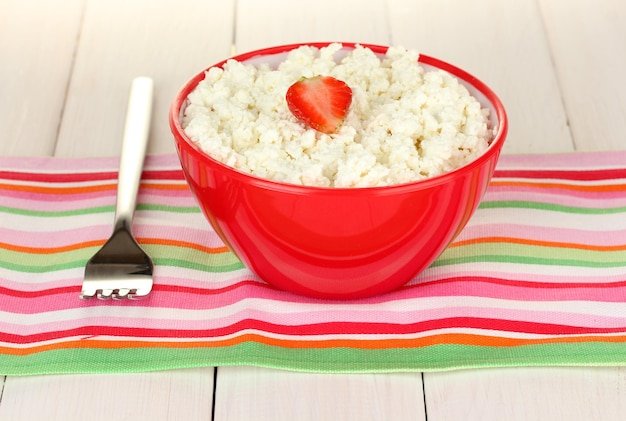 Cottage cheese with strawberry in red bowl and fork on colorful napkin on white wooden table closeup