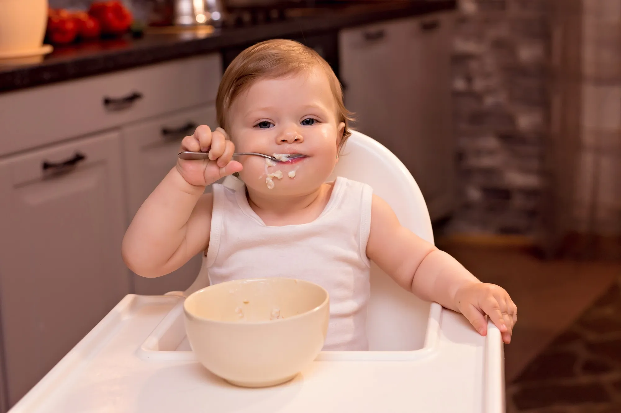happy child eating milk porridge with spoon, girl sitting on feeding chair in kitchen