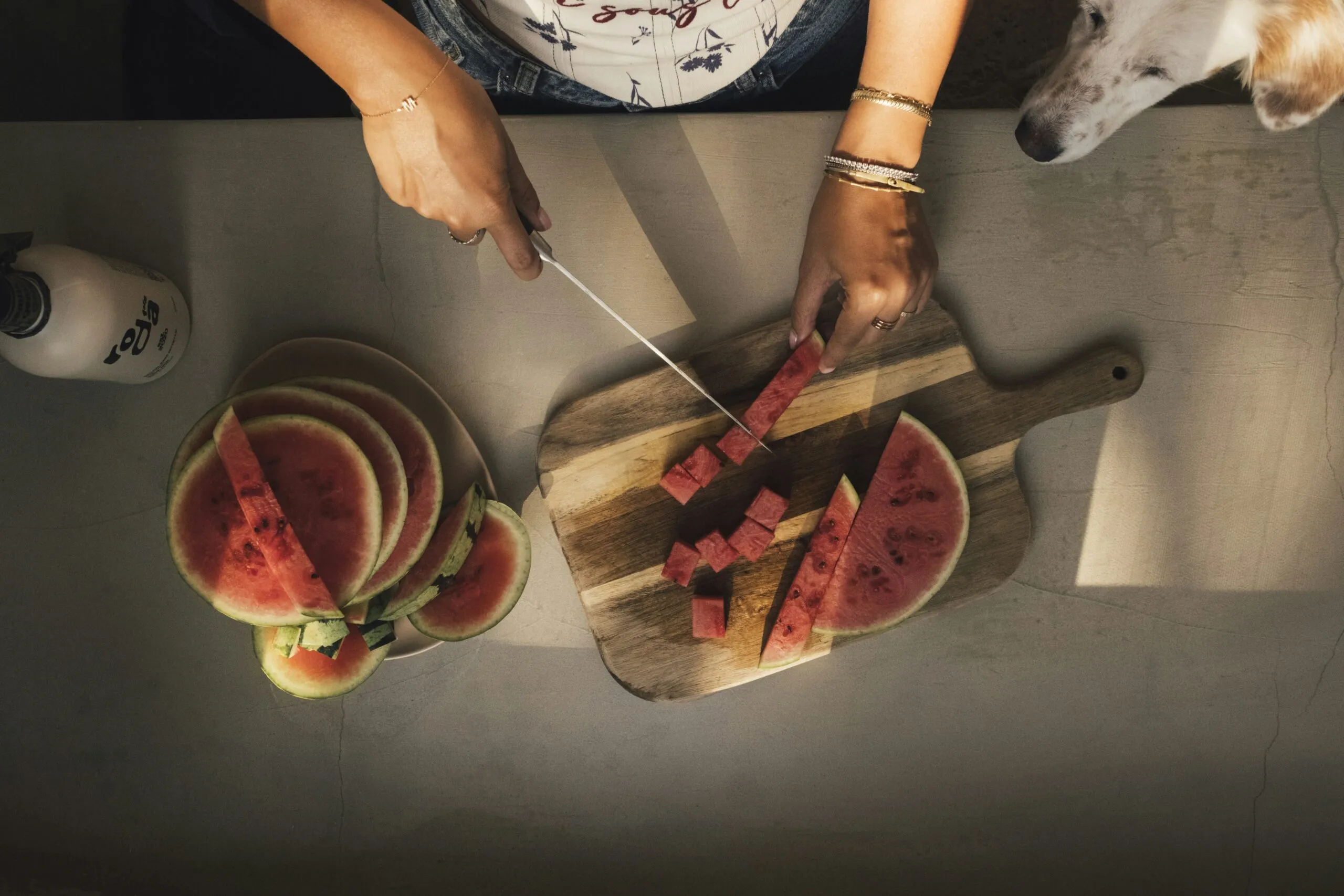 Hands slicing watermelon on a cutting board, dog watches.