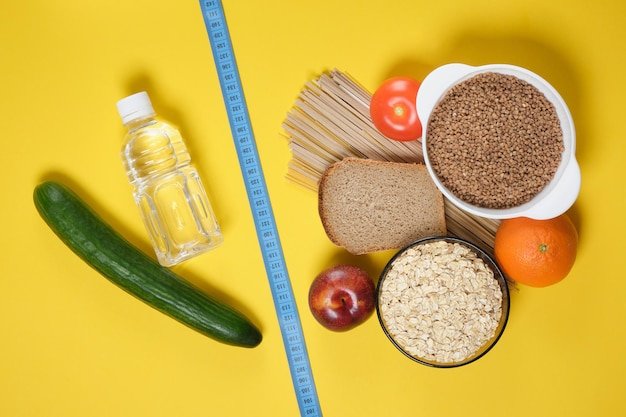 Measure tape, fruits, vegetables, cereals, bread, noodles and a bottle of water on a yellow background copy space top view diet concept