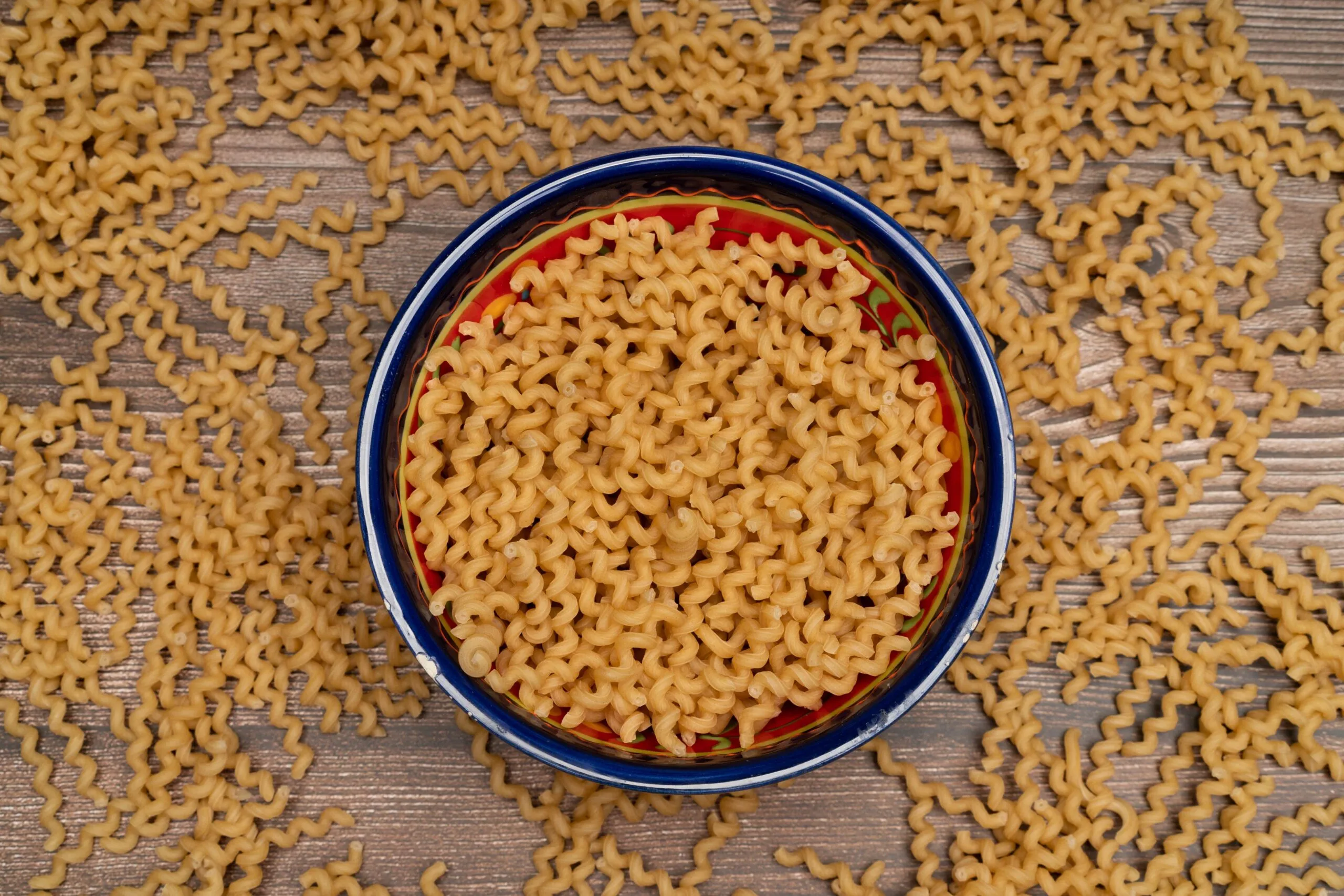 Top view of a ceramic bowl filled with uncooked fusilli pasta on a wooden surface, creating a still life composition.