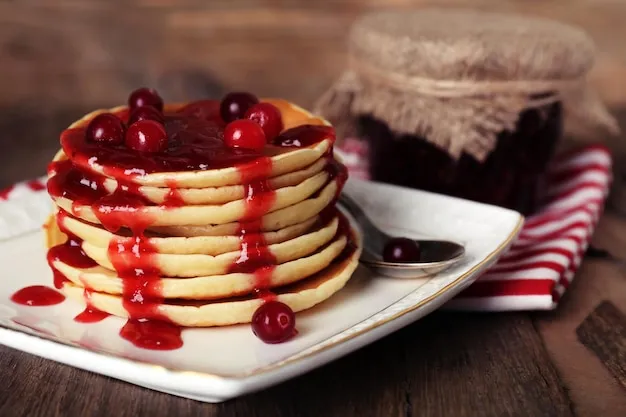 Stack of delicious pancakes with cranberries and jam on plate and napkin on wooden background