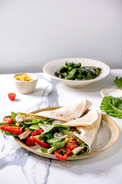 Traditional mexican tortila wrap with pork meat and vegetables in ceramic plate served with grilled green peppers jalapenos and corn over white marble table. Homemade fast food