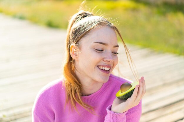 Young pretty girl at outdoors holding an avocado with happy expression