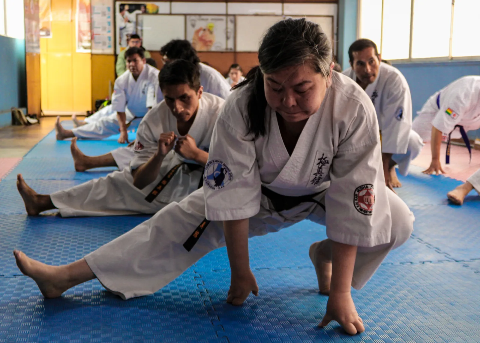 Sensei Akemi Watanabe Sosaki (2nd Dan) leads a training session in which Kyokushin-Kan Karate practitioners perform stretching exercises focused on hip opening and leg flexibility as part of their physical conditioning routine. The image shows the Sensei and her students wearing their dogi while carrying out technical preparation exercises on the tatami of the dojo in the city of La Paz, Bolivia.