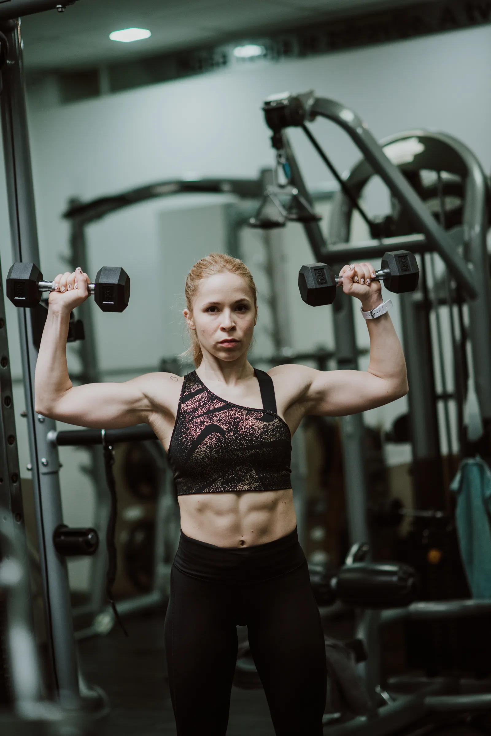 A determined woman lifts dumbbells overhead in a fitness gym. She showcases her strength and dedication while focusing on her workout. The gym is equipped with various exercise machines.