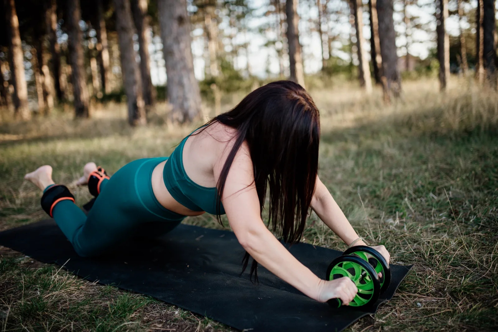 Woman doing exercises with an ab wheel and ankle weights on a yoga mat in the park.