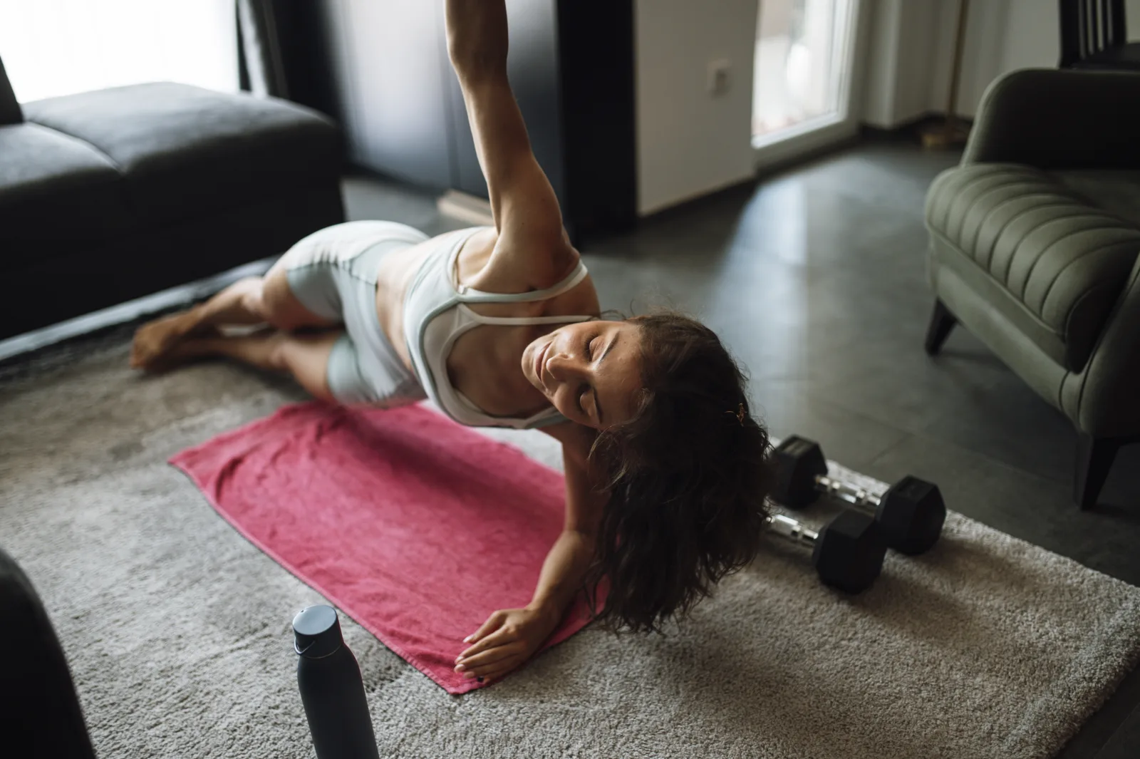 A woman performs a side plank on a pink mat in her living room. Dumbbells are placed nearby and sunlight fills the space through large windows.