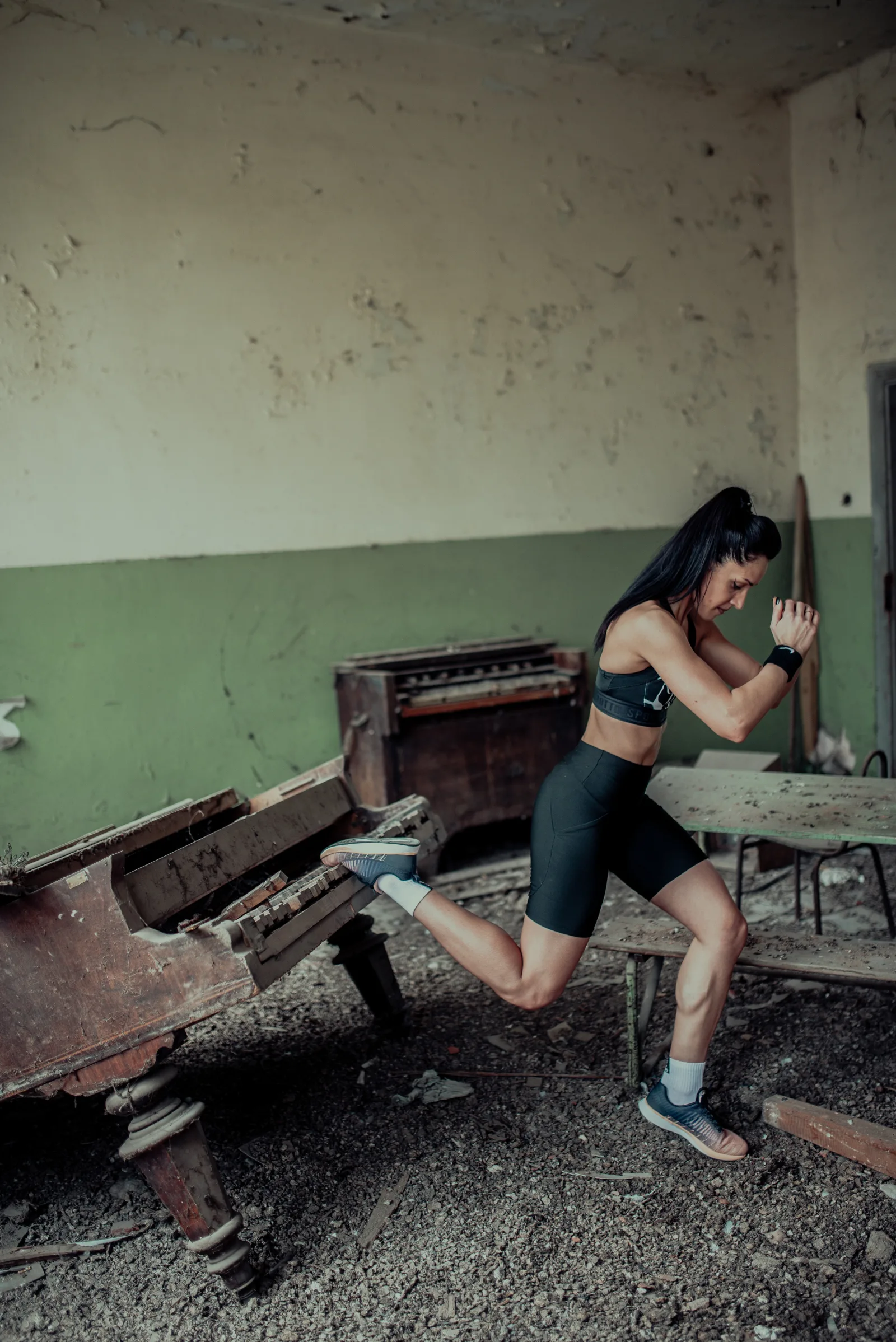 A woman performs a dynamic lunge while balancing on one leg in an abandoned building. The setting features old furniture and peeling walls that enhance the workout's intensity.