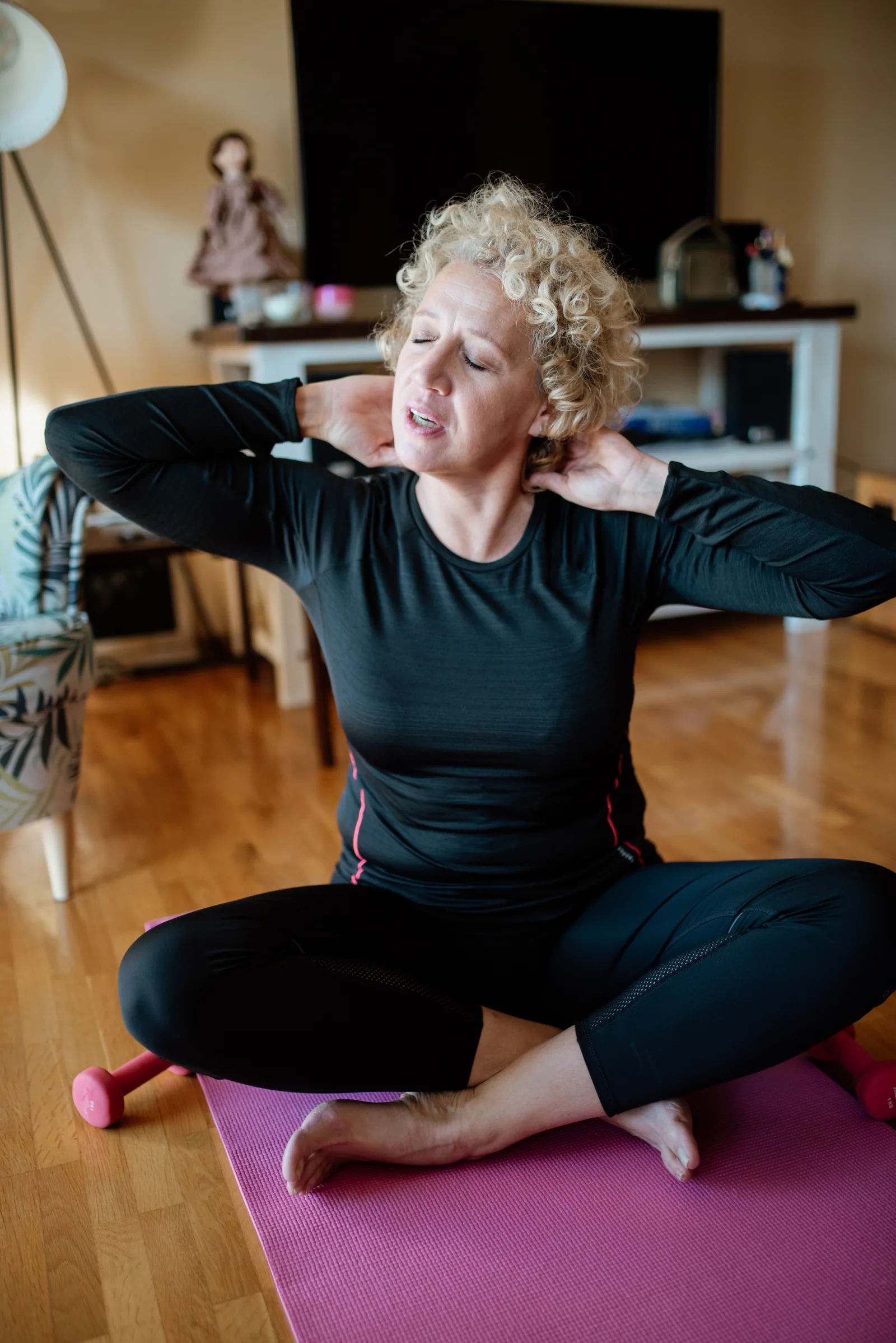 A woman sits on a mat in her living room, stretching her neck while seated cross-legged. She looks focused on finding comfort. It is a time for personal care and wellness.