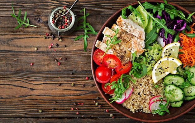 Healthy dinner. Buddha bowl lunch with grilled chicken and quinoa, tomato, guacamole, carrot, red cabbage, cucumber and arugula on wooden table. Flat lay. Top view