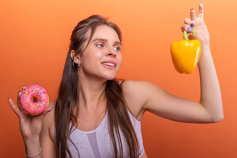 Woman in sports bra juggling choice between a donut and bell pepper on orange background.