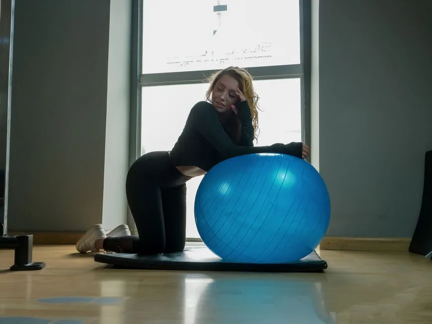 A woman in sportswear kneeling on a mat with a blue fitness ball indoors.