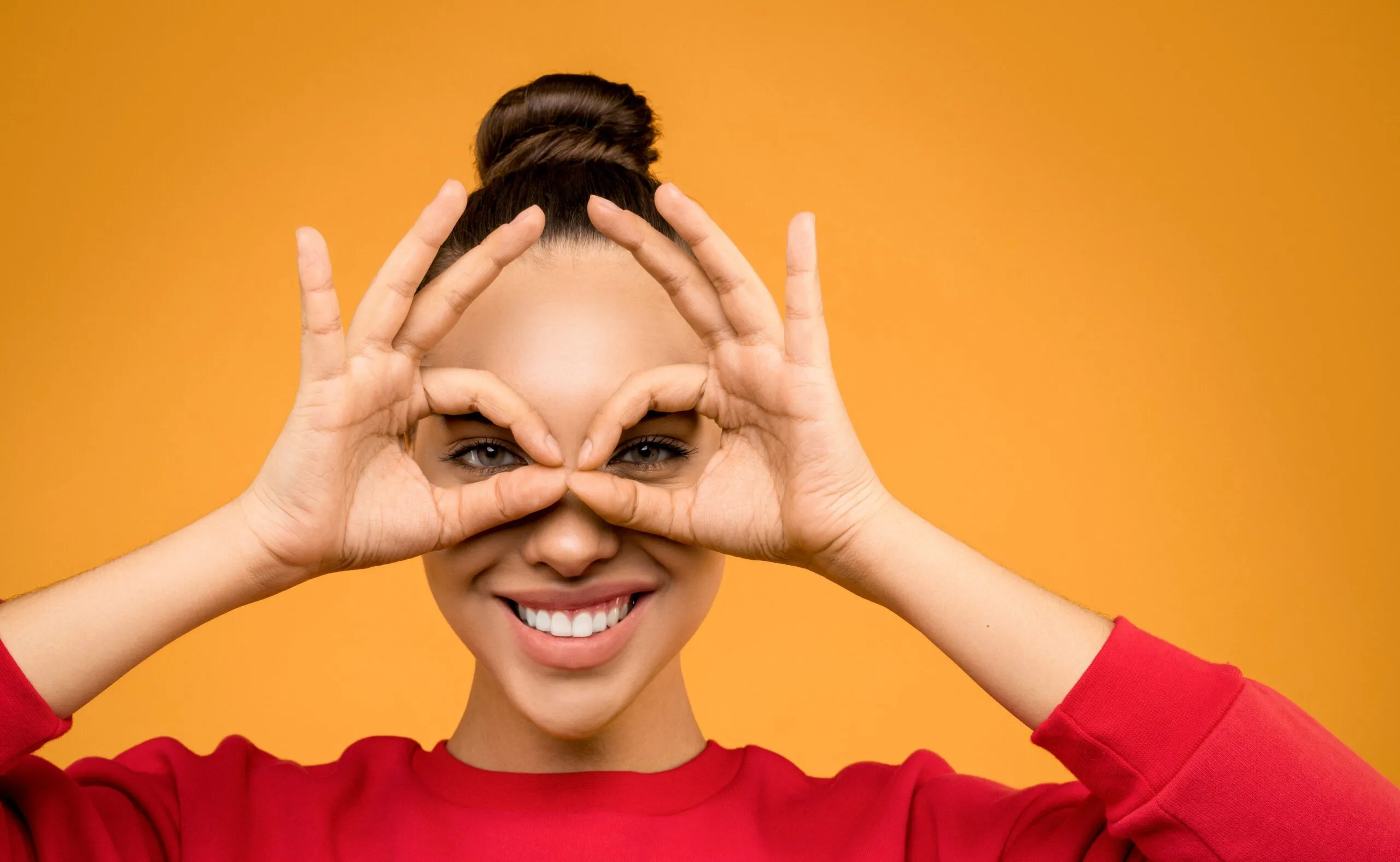Portrait of a smiling woman making a playful eye gesture with her hands against an orange background.