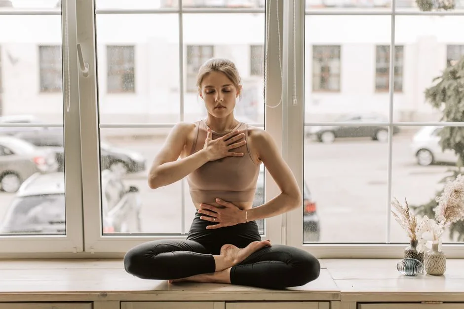 A woman practicing meditation in a serene yoga pose by a large window.
