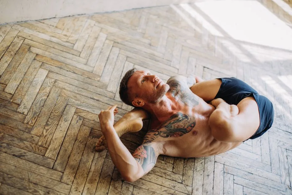 Tattooed man performing advanced yoga stretch indoors on wooden floor.