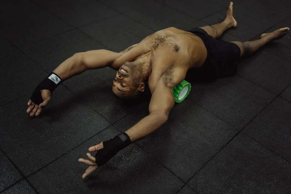 Topless man using foam roller for muscle recovery in gym setting.