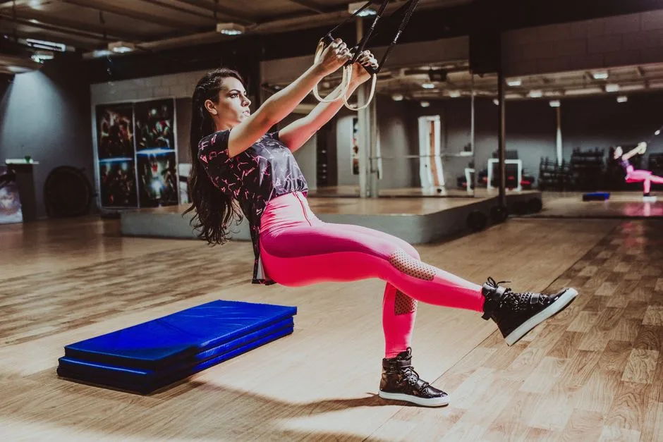 Full length focused young sportswoman in pink leggings squatting on one leg holding TRX straps while working out in equipped fitness center