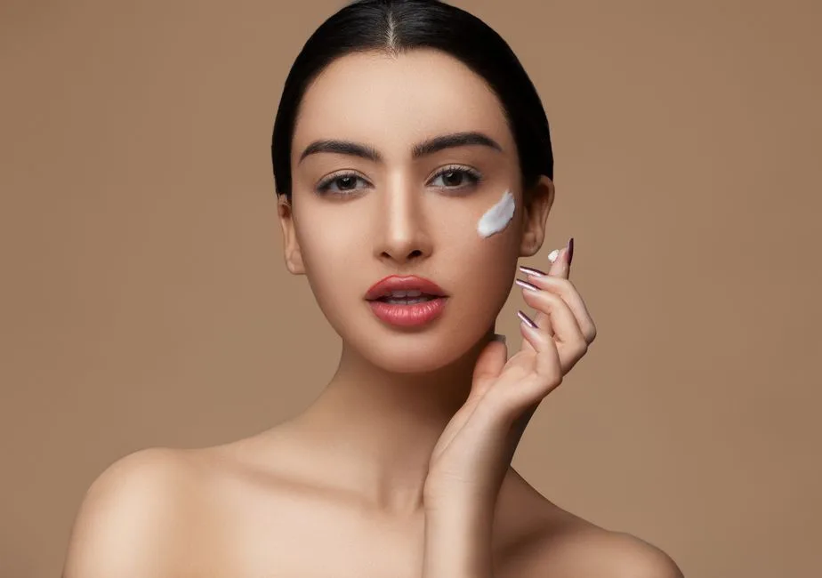 Crop sensitive female with makeup and red lips applying cream on delicate face skin while looking at camera on beige background