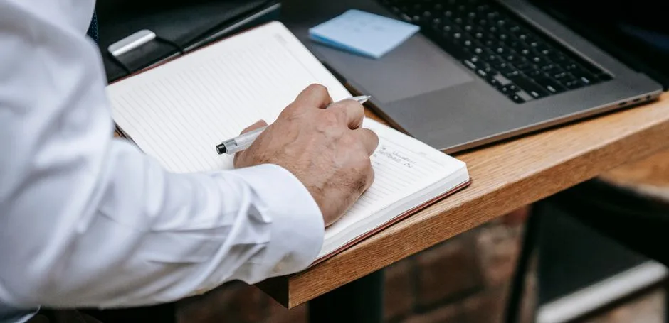 Close-up of an adult writing in a notebook beside a laptop. Professional setting.