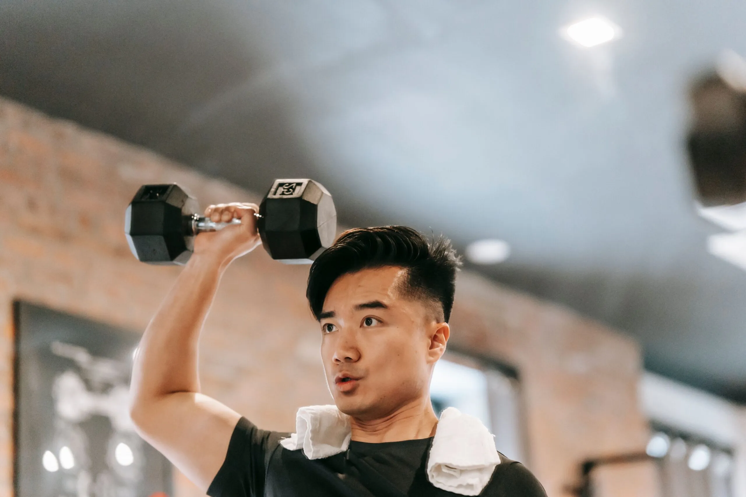 Powerful Asian male with towel standing with raised arm and lifting heavy dumbbell while pumping bicep in spacious gym on blurred background