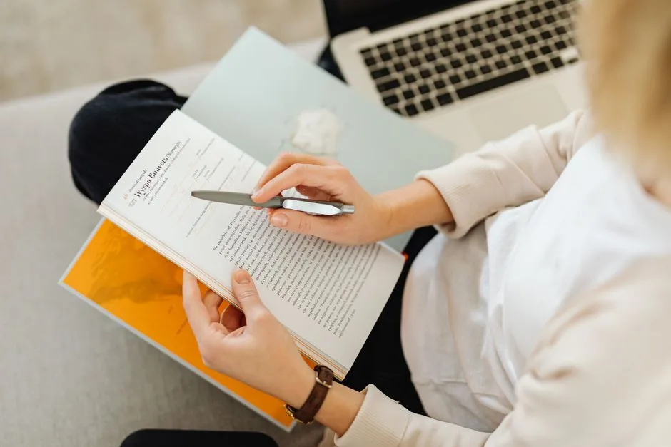 A person in white sweater reading a book with a pen and laptop in view.