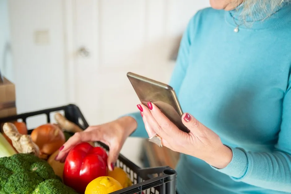 Woman in a blue sweater using a smartphone while shopping for vegetables indoors.