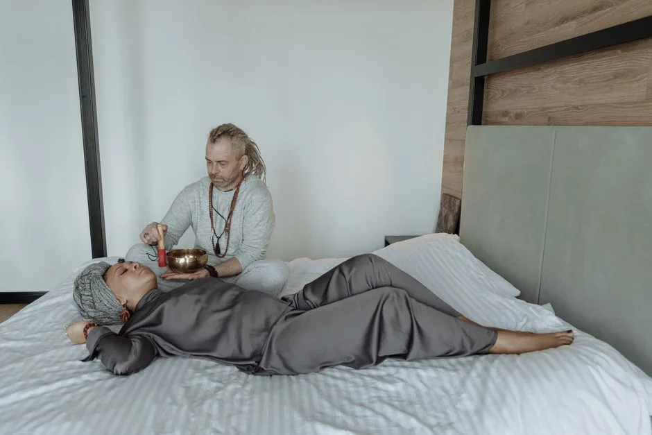 A man uses a singing bowl for sound therapy on a resting woman, promoting relaxation and spiritual healing.