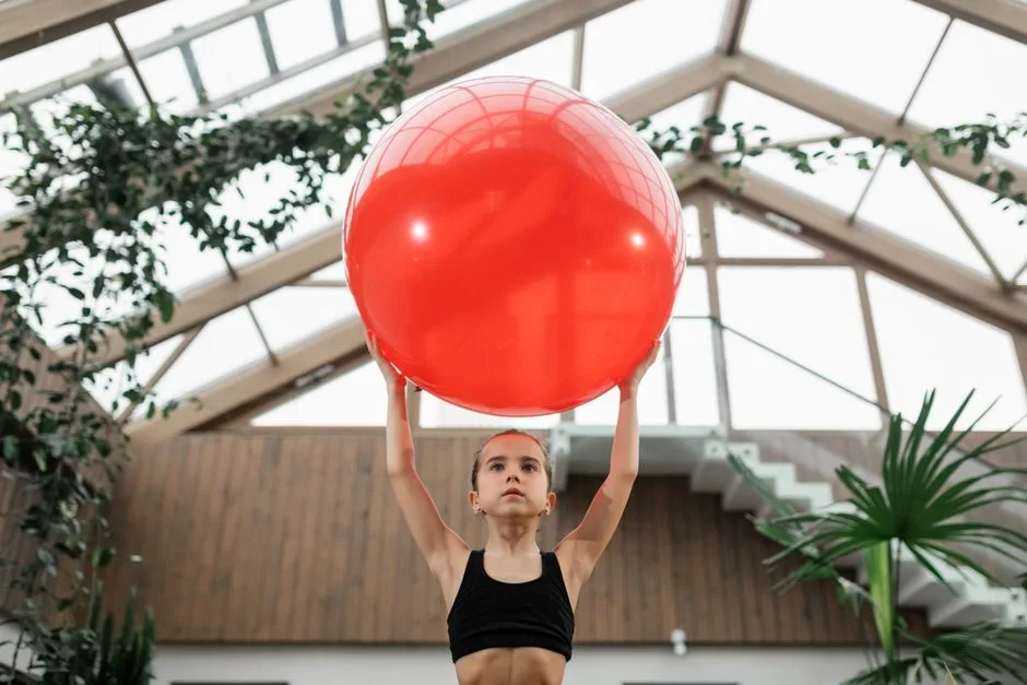 Young girl exercising with a red fitness ball in a stylish indoor gym surrounded by plants.