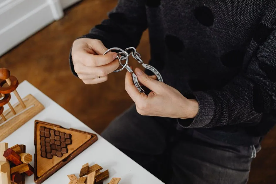 Top view of hands solving metal puzzle, surrounded by wooden brain teasers on a table.