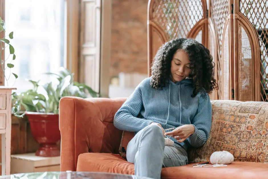 Young woman knitting on a couch in a warm, cozy indoor setting, wearing a blue sweater.