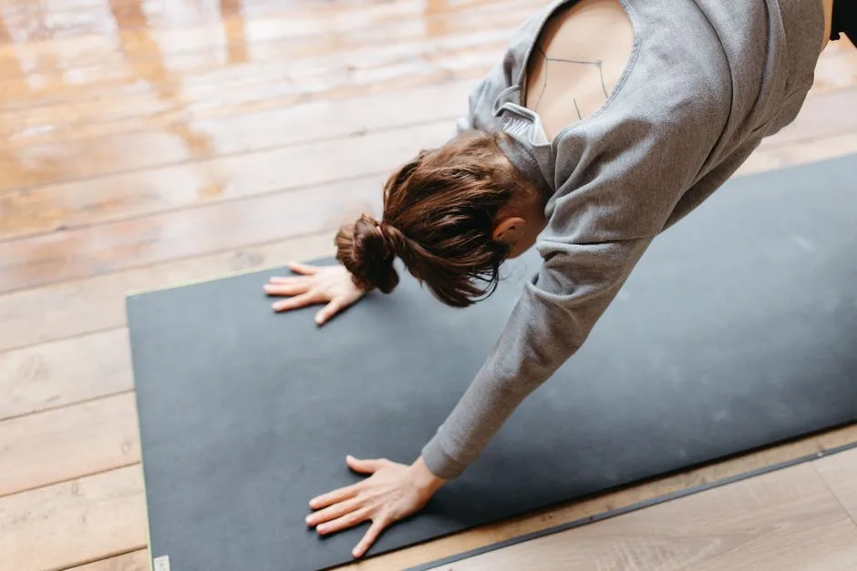 Young woman in gray sweatshirt doing yoga on a mat indoors on a wooden floor.