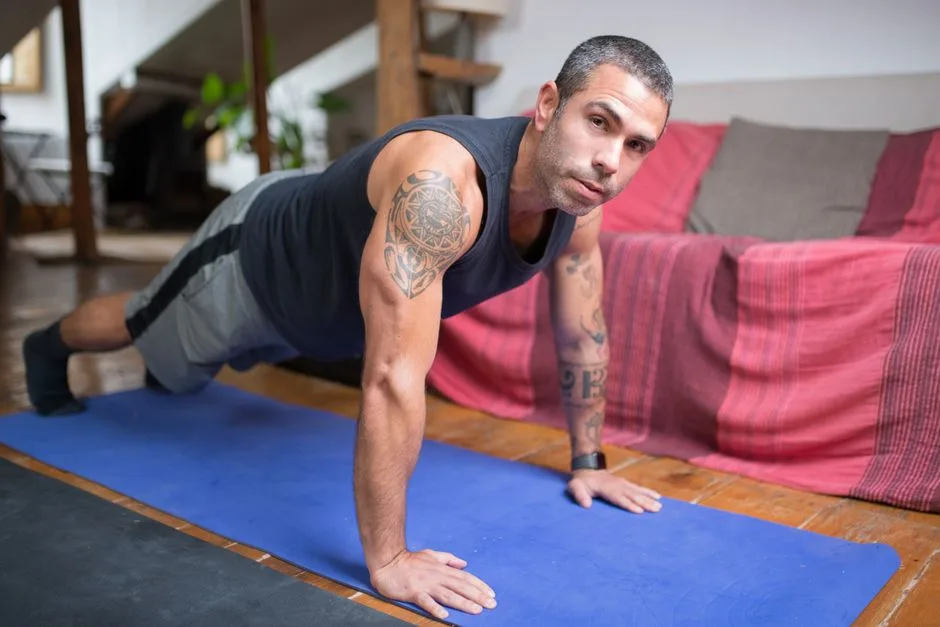 A tattooed man performs push-ups on a yoga mat indoors, focusing on fitness training.