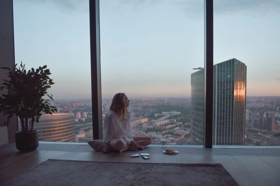 A woman sits by a large window, enjoying the sunset view of skyscrapers in a modern city.