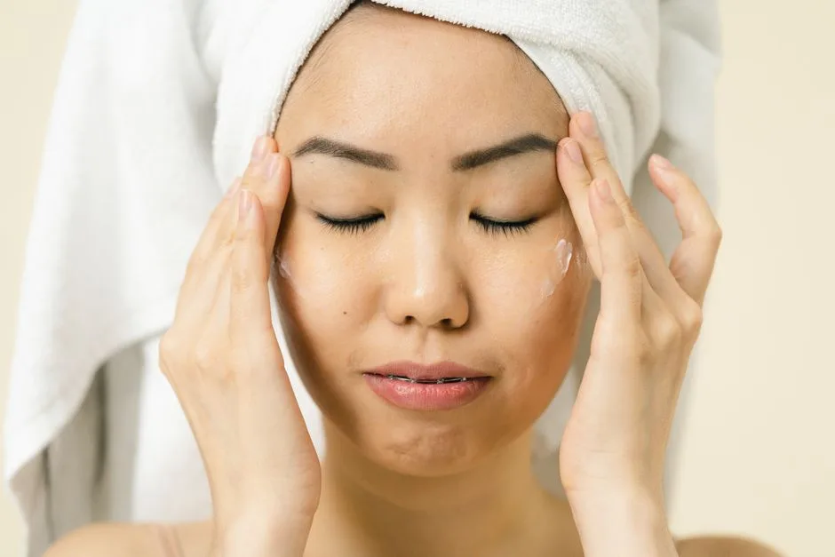 Asian woman with a towel on head applying moisturizer for skincare routine.