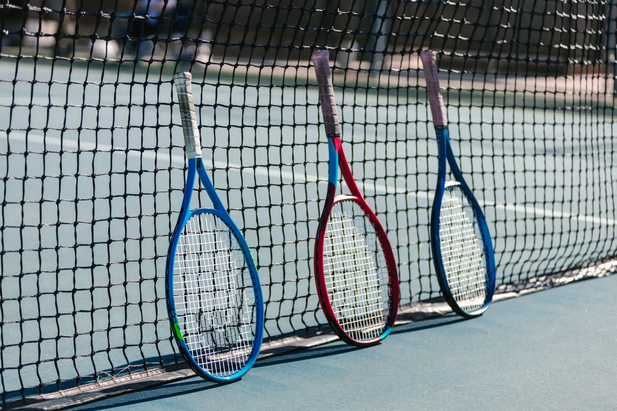 Three colorful tennis rackets leaning against a net on an outdoor tennis court.