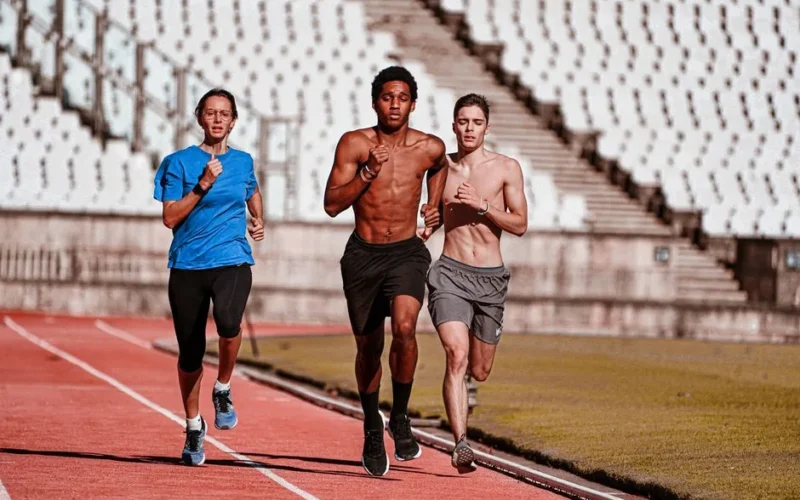 Three people running on a red track in a stadium, showcasing teamwork and physical fitness.