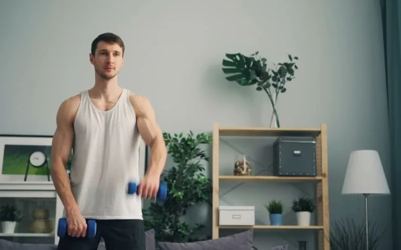 A muscular man in a tank top exercises with dumbbells indoors during the day.