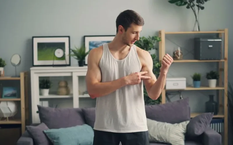 Muscular man in a tank top standing indoors, examining his biceps with a modern living room background.