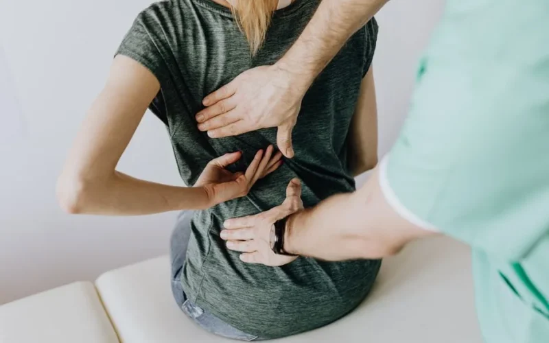 From above of crop faceless slim female patient sitting on examination couch and indicating osteopath in uniform pain spot on back in clinic