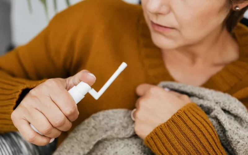 Adult woman using a mouth spray for relief indoors, wrapped in a cozy sweater.